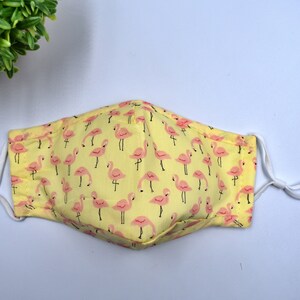 May include: A yellow face mask with a pink flamingo pattern. The mask has white elastic ear loops.