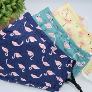 May include: Three reusable face masks with a flamingo print. The masks are navy blue, teal, and yellow with pink flamingos.