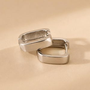 May include: A pair of silver-toned hoop earrings with a rectangular shape. The earrings feature a smooth, polished metallic finish and a hinged closure mechanism, designed as small, thick hoops with rounded corners and a hollow center.
