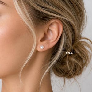 May include: A single round white pearl stud earring worn in the earlobe. The pearl has a smooth, lustrous surface and is positioned centrally. The text 6mm is visible next to the earring, indicating the diameter of the pearl stud in millimeters.