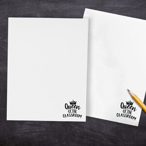 May include: Two white sheets of paper with the text "Queen of the Classroom" printed on each. A yellow pencil is on the right sheet of paper.