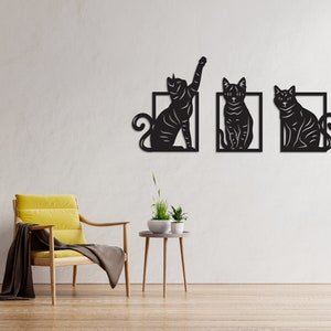 以下が含まれることがあります： 異なるポーズの猫のシルエットが描かれた、黒い金属製の壁飾り3点。各猫は正方形の枠で囲まれています。