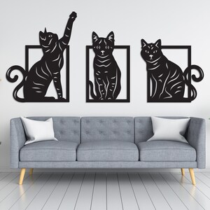 以下が含まれることがあります： 異なるポーズの猫のシルエットが描かれた、黒い金属製の壁飾り3点。各猫は正方形で囲まれています。