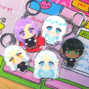 Puede incluir: Cinco llaveros de acrílico con personajes estilo chibi de distintos colores de pelo, incluyendo morado, blanco y negro. Cada personaje viste un atuendo oscuro o claro con un pequeño emblema de calavera. Incluyen anillas y cadenas de metal.