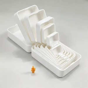 May include: A set of nested white rectangular plastic containers arranged in a cascading formation, with the largest at the back and smallest at the front. A small, orange, hand-shaped figurine displaying a middle finger gesture sits in front.