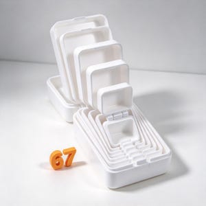 May include: A set of white plastic rectangular storage containers of varying sizes, nested together in a tiered arrangement. The containers feature hinged lids, and an orange plastic number 67 is positioned to the left of the nested set.