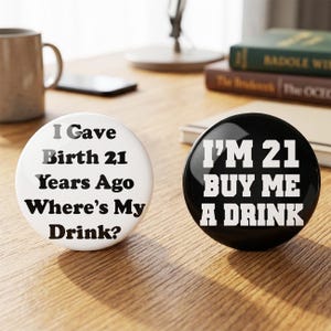 May include: Two circular pin-back buttons. The left button is white with black text that reads: I Gave Birth 21 Years Ago Where's My Drink? The right button is black with white text that reads: I'M 21 BUY ME A DRINK. Both buttons are round.