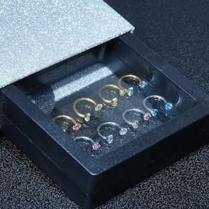 May include: A collection of horseshoe-shaped nose rings in a black box. The rings are gold and silver-toned, each adorned with a colorful gemstone. The box is set against a sparkly silver surface.