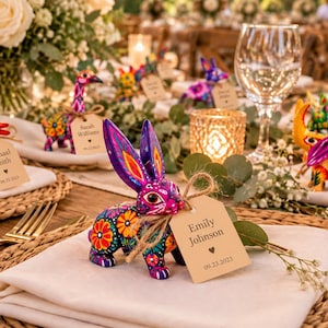 May include: A hand-painted purple rabbit figurine with intricate floral patterns in orange, yellow, and green. A tan rectangular paper tag tied with twine hangs from its neck, displaying the text Emily Johnson and 09.23.2023.