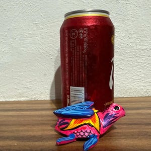 May include: A small, hand-painted wooden turtle figurine featuring vibrant pink, blue, and yellow patterns, positioned in front of a red aluminum beverage can with white text and a barcode visible on its side.