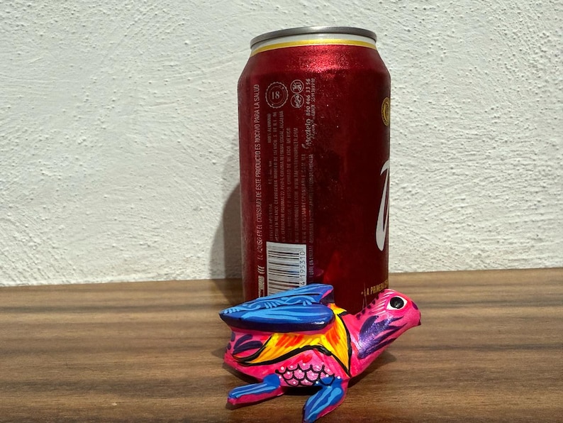 May include: A small, hand-painted wooden turtle figurine featuring vibrant pink, blue, and yellow patterns, positioned in front of a red aluminum beverage can with white text and a barcode visible on its side.