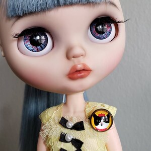 May include: A doll with blue hair and red and blue eyes wearing a yellow dress with black bows and a cat pin.