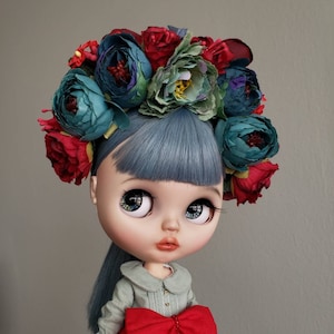 May include: A doll with blue hair and large blue eyes wears a flower crown with red, green, and blue flowers. The doll is wearing a light blue shirt with a white collar and a red bow.