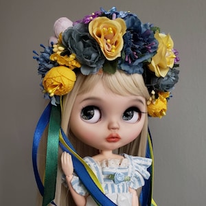 Può includere: Una bambola con i capelli biondi che indossa una corona di fiori blu e gialli e un vestito a righe blu e bianche. La bambola ha grandi occhi blu e un viso delicato.