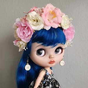 May include: A blue-haired doll wearing a floral dress and a flower crown. The doll has large blue eyes and a pink blush on her cheeks.