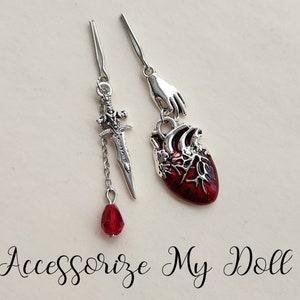 May include: Two silver charm earrings, one with a red heart and the other with a silver sword and a red teardrop charm. The earrings are on a white background with the text "Accessorize My Doll".
