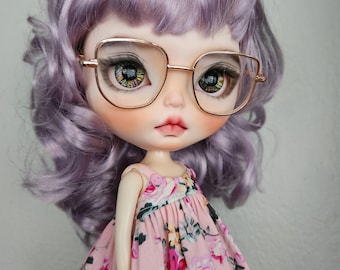 May include: A doll with wavy lavender hair and large, detailed eyes wearing gold-rimmed glasses. The doll has a pale complexion, pink lips, and is dressed in a sleeveless light pink dress featuring a vibrant floral pattern with roses.