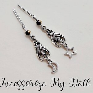 May include: Silver-toned dangle earrings featuring a bat-like design with a skull, a star, and a crescent moon charm. The earrings have a dark bead accent and are displayed on a white surface. The text "Accessorize My Doll" is at the bottom.