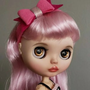 Op de afbeelding: Een roze pop met lang roze haar en een roze strik hoofdband. De pop heeft grote bruine ogen en draagt een roze en zwarte jurk.