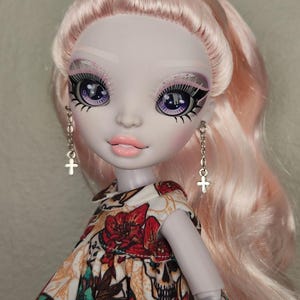 May include: A doll with pale skin and long, pink hair. She is wearing a white dress with a floral and skull pattern. She has purple eyeshadow and blue eyes.
