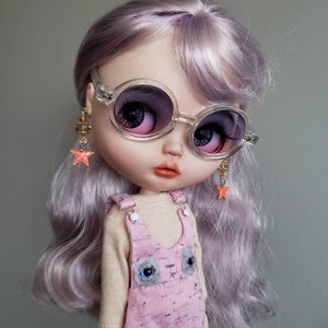 May include: A doll with long, wavy, lavender hair wearing clear round sunglasses and pink overalls with a cat face design. The doll is wearing star-shaped earrings.