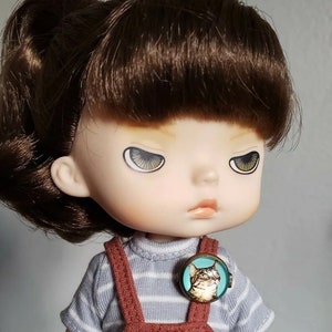 May include: A doll with brown hair in pigtails, wearing a striped shirt and red overalls. The doll has a serious expression and is wearing a round pin with a cat on it.