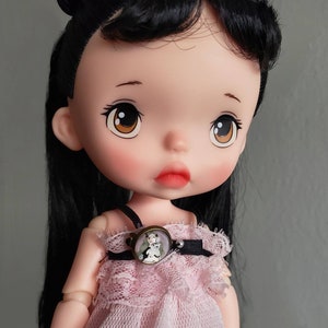 May include: A doll with black hair and large brown eyes is wearing a pink dress with lace trim and a necklace with a cameo pendant.