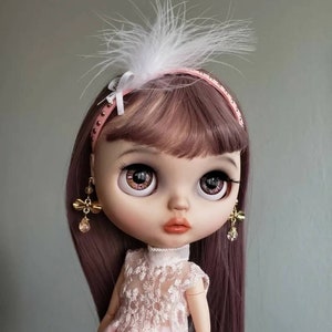 Op de afbeelding: Een pop met lang paars haar, die een roze hoofdband met een witte veer, een roze jurk met kant en gouden oorbellen met een strik draagt. De pop heeft grote blauwe ogen met roze glitter.