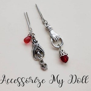 May include: Two silver hair clips with red glass beads and charms. One charm is a bat with a skull, the other is a hand with a ring.  Accessorize My Doll.
