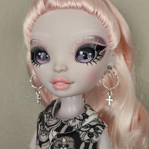 May include: A doll with pale skin, pink hair, and purple eyes. She is wearing a black and white dress with a cat and dog print. She has silver hoop earrings with a cross charm.