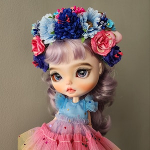 May include: A doll with purple hair wearing a blue and pink dress and a flower crown with blue, pink, and white flowers.