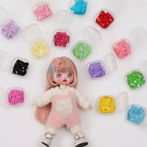May include: A doll with pink hair and a pink and white outfit is surrounded by small containers of colorful hair clips. The containers are clear plastic and each contains a different color of hair clips.