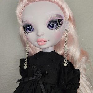 May include: A doll with pale skin, pink hair, and purple eyes. She is wearing a black dress with a black rose on the front and silver skull earrings.