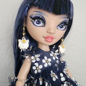 May include: A doll with long, dark blue and black hair, wearing a navy blue dress with white flowers. The doll has blue eyes and is wearing gold earrings with a white cat charm.