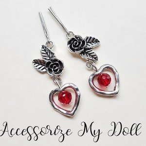 May include: A pair of silver earrings with a red heart charm and a rose and leaf design.