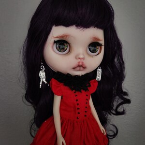 May include: A doll with long, dark purple hair, wearing a red dress with black lace trim and black accents. The doll has large, green eyes and is wearing silver earrings with charms.