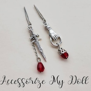 May include: Two silver earrings with red gemstone drops. One earring features a silver sword charm and the other features a silver hand charm.