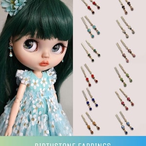 May include: A doll wearing a blue dress with white flowers and a green flower in her hair. The doll is wearing earrings with different coloured crystals. The text "BIRTHSTONE EARRINGS" is at the bottom of the image.