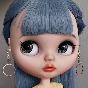 Può includere: Una bambola con i capelli blu e grandi occhi azzurri. La bambola indossa orecchini a cerchio dorati.