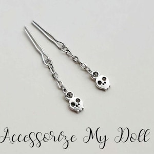 May include: Two silver skull hair clips with a chain connecting the skull to the clip. The clips are on a white background.