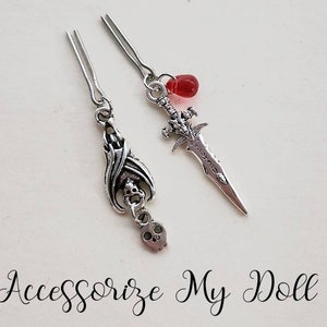 May include: Two silver hair clips with charms. One clip has a skull and bat charm, the other has a sword charm with a red bead.