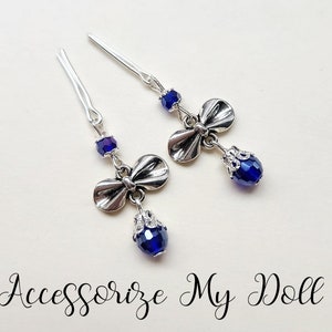 Op de afbeelding: Twee zilveren haarspelden met een strik en een blauwe kristallen kraal. De spelden hebben een decoratieve strik charme en een druppelvormige blauwe kristal. De tekst "Accessorize My Doll" staat onderaan de afbeelding.