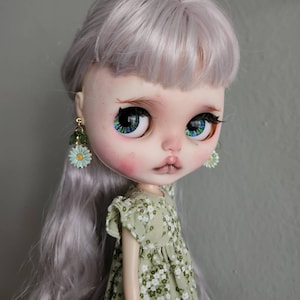 May include: A doll with long, silver hair wearing a green floral dress and blue and green flower earrings.