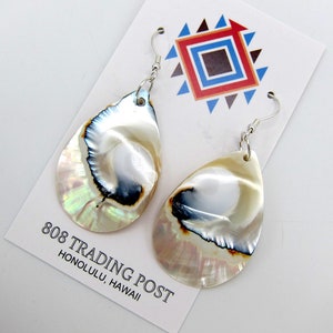 May include: A pair of iridescent white shell earrings with a black and brown pattern. The earrings are hanging from silver hooks and are displayed on a white card with the text "808 TRADING POST HONOLULU, HAWAII".