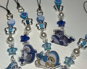Puede incluir: Una colección de colgantes para teléfono con cuentas. Cada colgante presenta un gato con un disfraz de tiburón, con detalles en azul y blanco. Los colgantes están ensartados con cuentas transparentes, perlas y detalles en forma de estrella, y están sujetos a cordones negros.