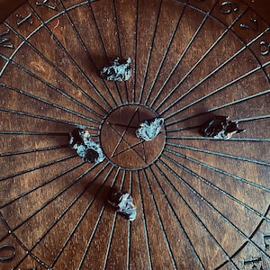 May include: A wooden Ouija board with a pentagram in the center and four black stones on the board. The board has letters, numbers, and words around the edge.