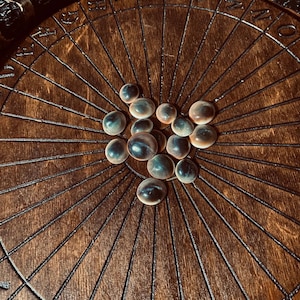 May include: A dark wooden divination board featuring a radial design and etched symbols. The board has a network of lines radiating from the centre. A collection of small, round, iridescent stones are arranged on the board, suggesting a spiritual or mystical theme.