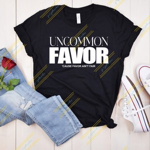 May include: Black t-shirt with white text that reads "Uncommon Favor 'Cause Favor Ain't Fair". The shirt is laid flat on a wooden surface with a pair of blue jeans and a red rose.