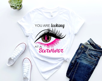 Könnte beinhalten: Weißes T-Shirt mit einem rosa Band und dem Text "You are looking at a Survivor" mit einer stilisierten Augengrafik.