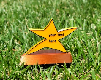 May include: A yellow star-shaped trophy with black trim and the words "your text here" on the front. The trophy sits on a brown base. The background is green grass.